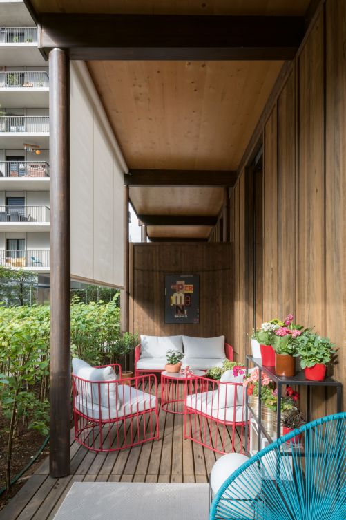 Von Frühling bis Herbst benutzbar: Die gedeckte Veranda mit Holzstützen ist der Lieblingsort der Familie und liegt direkt vor der Küche. Ana und Antonio möblierten sie mit farbenfrohen Gartenmöbeln von Giardimo LAB. Auf dem Metallregal gedeihen Blumen und Kräuter, eine Hecke trennt den privaten Bereich vom öffentlichen Weg.